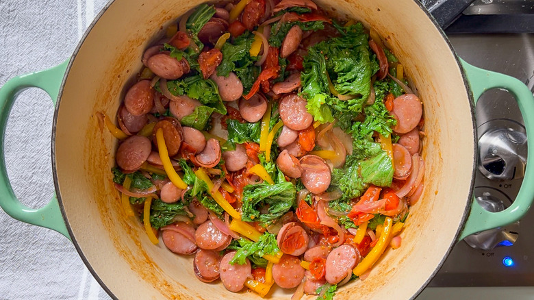 Sliced kielbasa, wilted mustard greens, tomatoes, yellow bell pepper and red onion in pot on stove top