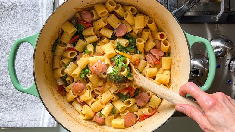Stirring cooked pasta and smoked mozzarella into mustard greens and kielbasa with wooden spoon on stove top.