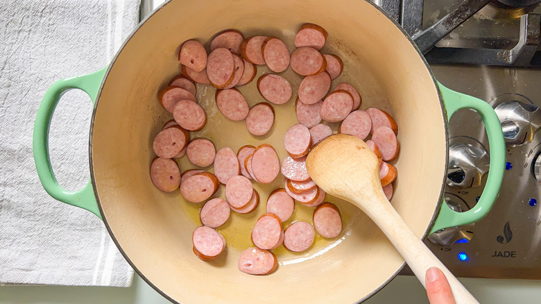 Stirring sliced kielbasa sausage in pot on stovetop with wooden spoon