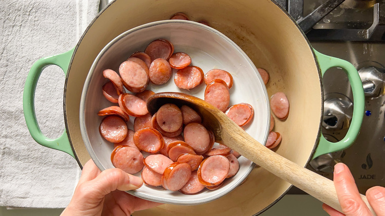 Transferring cooked sliced kielbasa from pot to plate with wooden spoon
