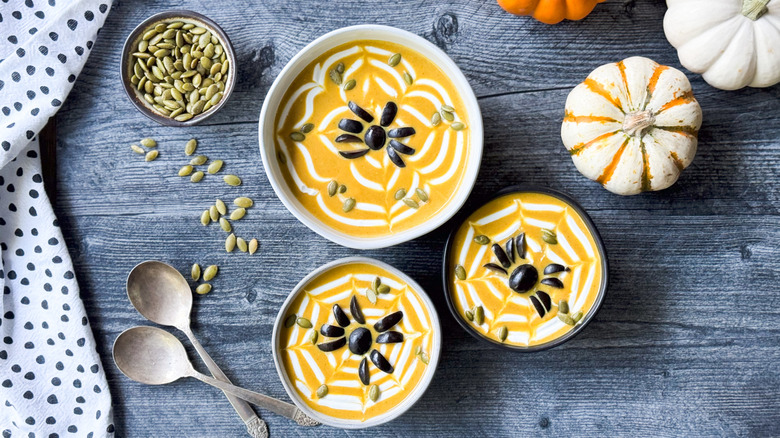 Spooky pumpkin curry Halloween soup in bowls with spiderweb sour cream, black olive spiders, and pumpkin seeds