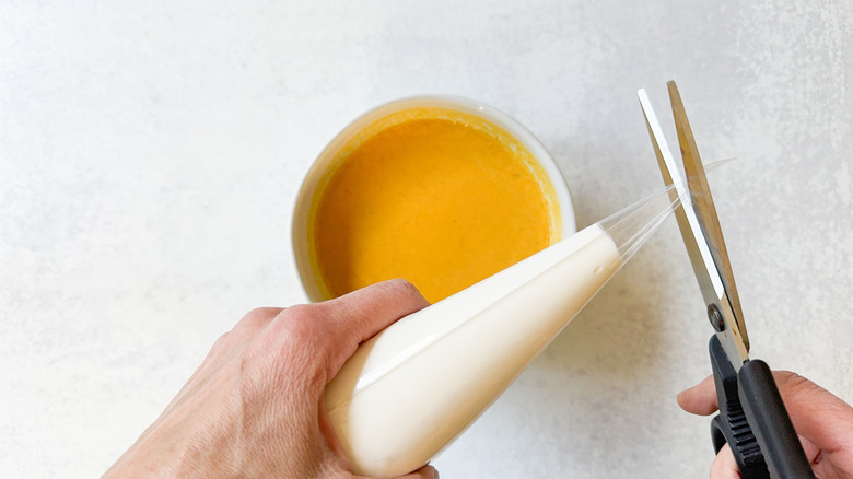 Cutting the tip off a disposable plastic pastry bag filled with sour cream and a bowl of pumpkin soup