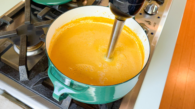 Blending spooky pumpkin curry Halloween soup with immersion blender in large pot on stovetop