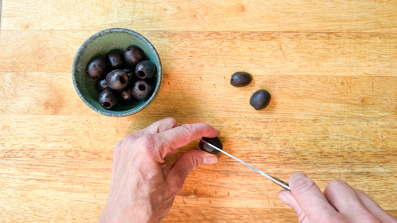 Cutting pitted black olives in half with small knife on cutting board