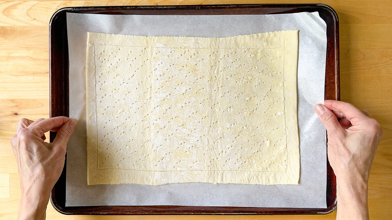 Lifting scored and pricked puff pastry on parchment paper onto a baking sheet