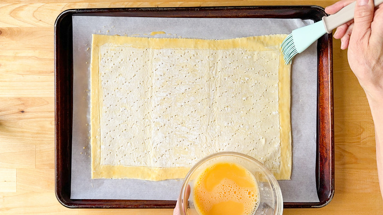 Using a pastry brush to apply egg wash to puff pastry on parchment paper on a baking sheet