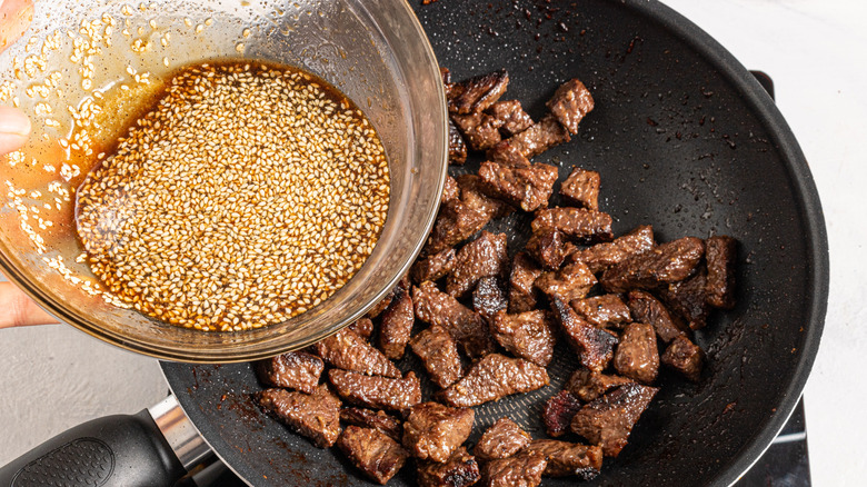 Adding soy sauce with sesame seeds to a wok with cooked meat pieces