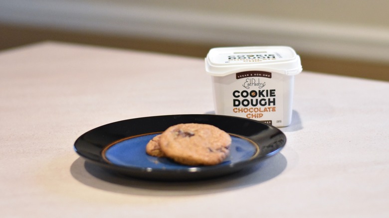 A container of Eat Pastry chocolate chip cookie dough with two baked cookies on a blue and black plate