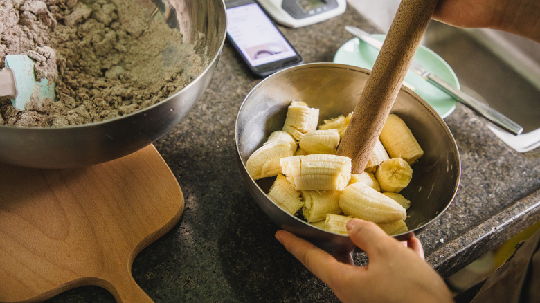 A person's hands mash bananas with a rolling pin in a metal bowl.