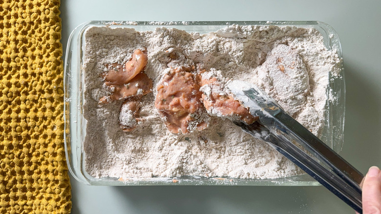 Dredging brined chicken thighs in flour mix in glass baking dish with tongs