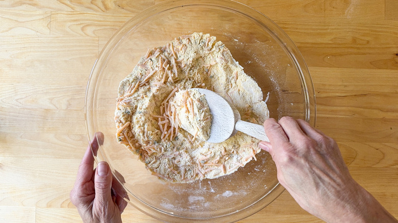 Mixing shredded cheddar cheese and thyme leaves into cornbread dry ingredients with wooden spoon in glass bowl