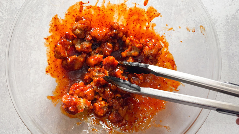 Tossing sweet and spicy Korean fried popcorn chicken with sauce in glass bowl with tongs