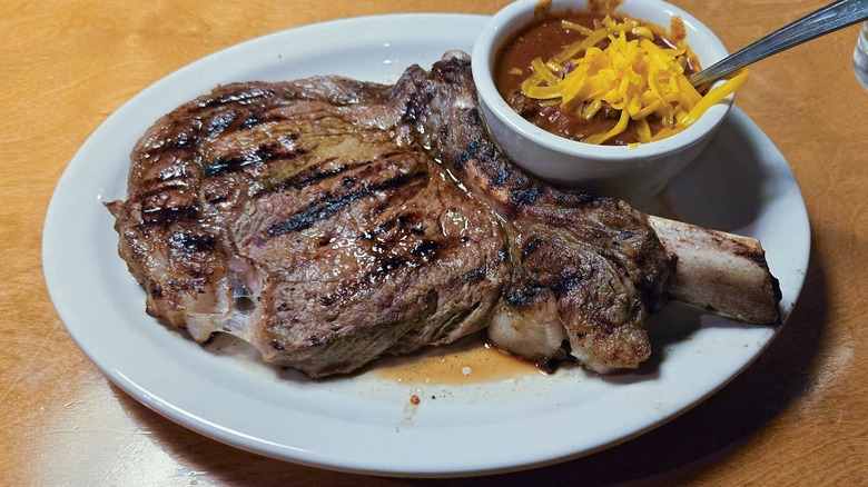 A bone-in ribeye steak on a white plate next to a bowl of chili