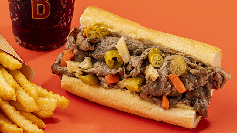 An Italian beef is on a table with fries and a drink.