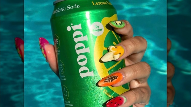 A woman with colorful painted nails holding a can of Lemon Lime poppi with water in the background