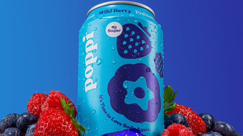 A Wild Berry poppi can of soda on a blue background surrounded by fresh berries