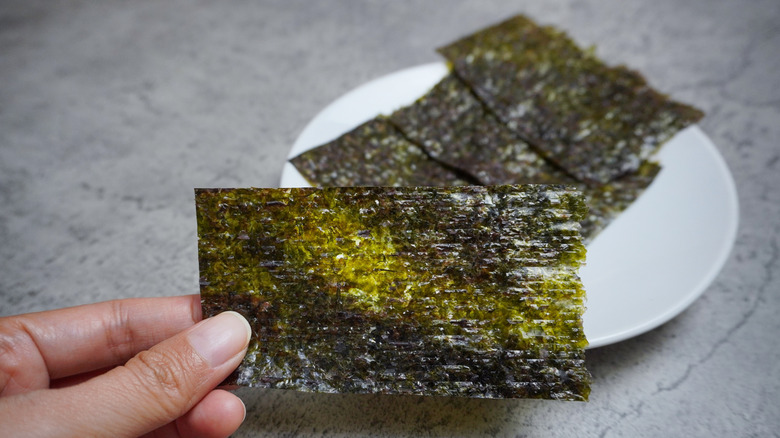 Hand holding seaweed sheet with plate of seaweed sheets in background