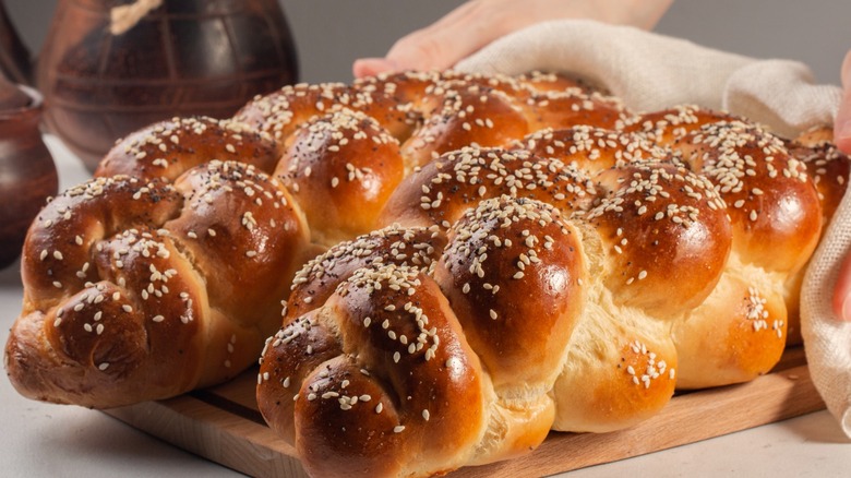 Golden-brown, sesame seed-covered braided challah loaves sit on a wooden cutting board