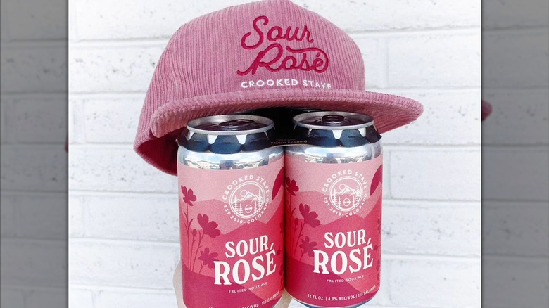 Two cans of beer with a pink hat on top with the name of the beer embroidered in dark pink