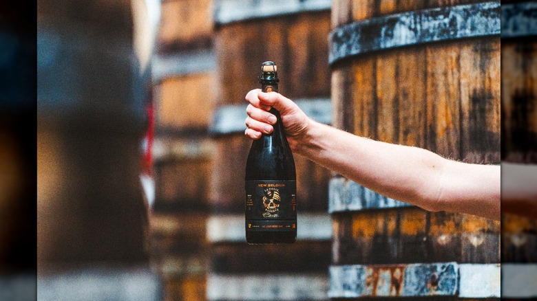 A woman holding a bottle of beer in one hand in front of wooden casks