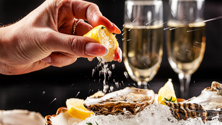 A hand squeezing lemon on fresh oysters with glasses of champagne in the background