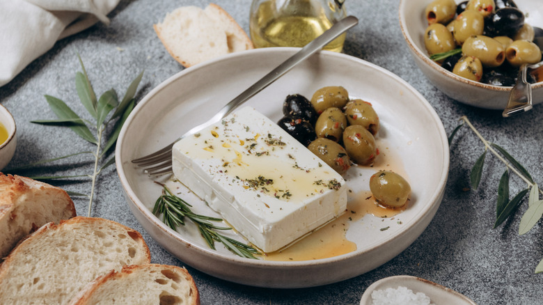 Feta cheese rests in a bowl beside some olives and fresh rosemary, drizzled with olive oil and herbs