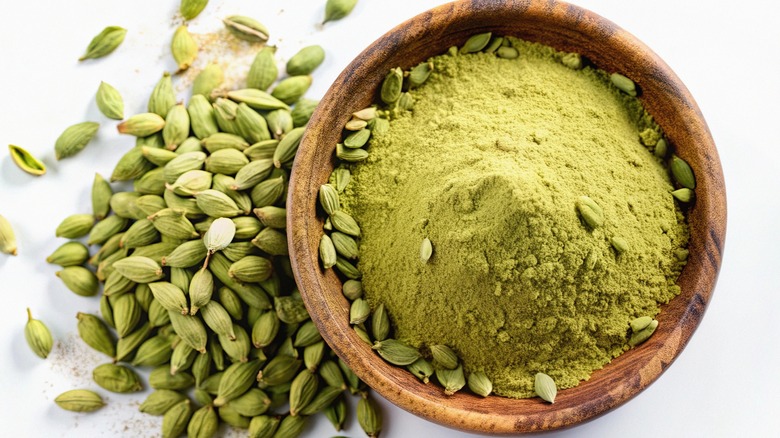 Ground cardamom in a wooden bowl surrounded by cardamom pods