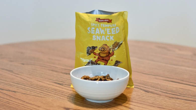 A bag of Trader Joe's Spicy Tempura Seaweed Snacks sits behind a white bowl of snacks on a wooden table.