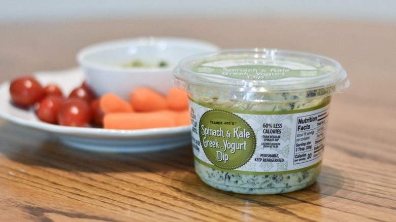 A package of Trader Joe's Greek yogurt dip sits in front of a white plate with vegetables on a wooden table.