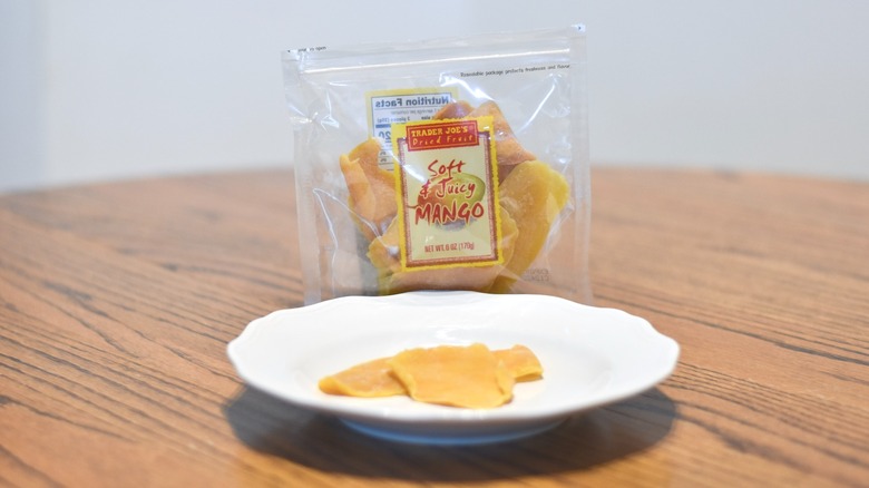 A bag of Trader Joe's dried mango has two slices on a white plate placed on a wooden table.