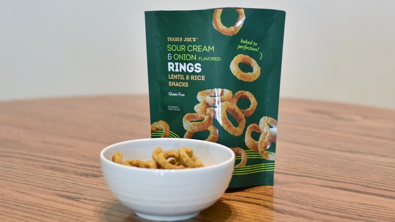 A bag of Trader Joe's Sour Cream and Onion Rings sits next to a white bowl of snacks on a wooden table.