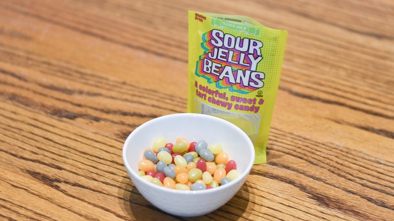 A bag of Trader Joe's sour jelly beans sits next to a white bowl of jelly beans on a wooden table.