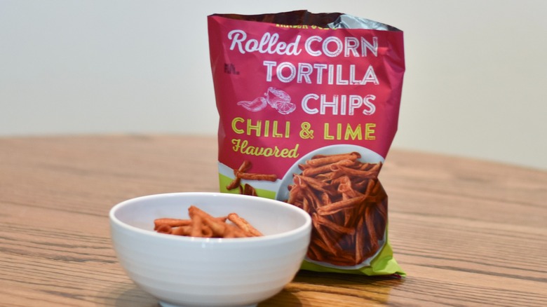 An open bag of Trader Joe's rolled corn tortilla chips has a white bowl of chips in front and sits on a wooden table.