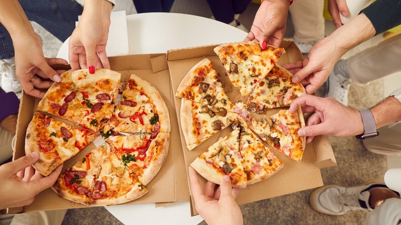 Various hands reaching for pizza slices in a delivery box