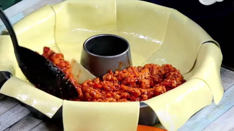 Black spoon placing meat over pasta sheets in a Bundt pan