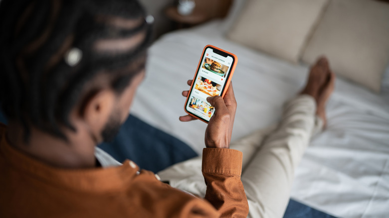 A man reclined on a bed ordering from a food delivery app on his phone