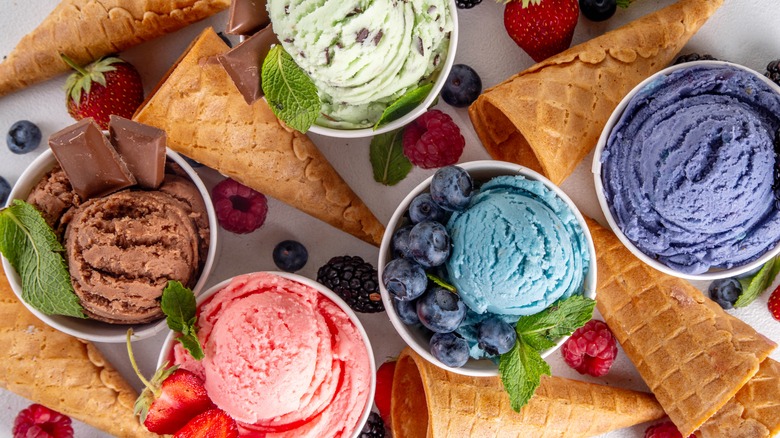 Cups of different flavors of ice cream on a white tabletop with fruit and waffle cones in between the cups