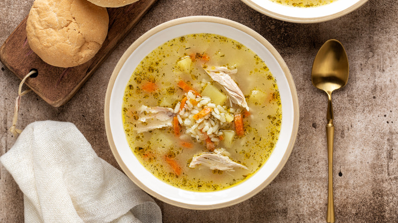 A bowl filled with chicken soup sitting in between a spoon and a napkin with a cutting board with rolls on it.