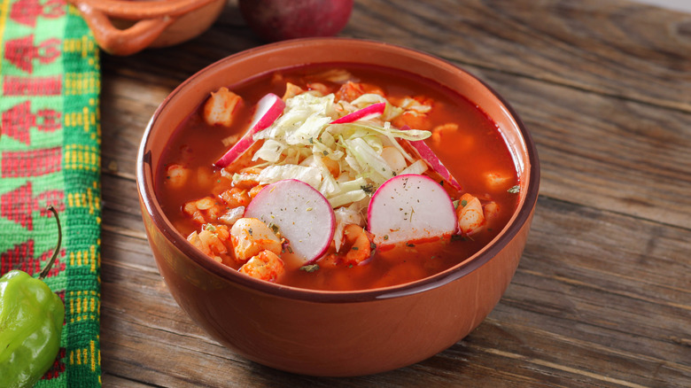 A clay bowl of traditional Mexican red pozole, topped with shredded lettuce and sliced radishes on a wooden table.