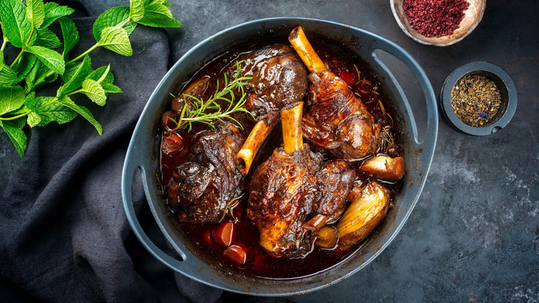 Roasted pork shank in a casserole dish surrounded by herbs and spices on a grey countertop.