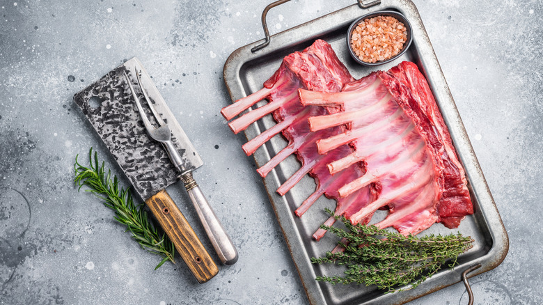 A metal tray containing two racks of lamb accompanied by seasoning and herbs.