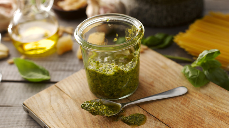 A glass jar of pesto sauce and a spoon of pesto sauce sitting on a wooden cutting board.