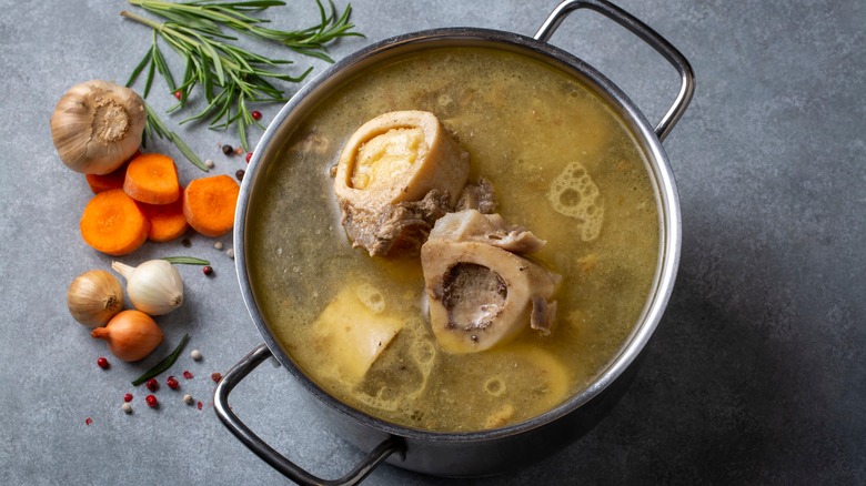 Pot of bone broth next to carrots, rosemary, garlic, onions, and peppercorn