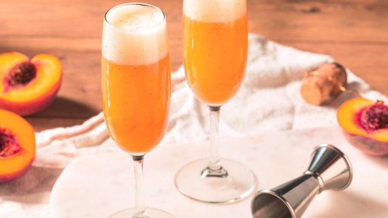 Two orange Bellini cocktails sit on a table near peach halves and a jigger.