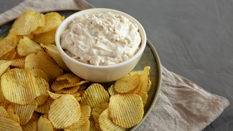 bowl of French onion dip sits on a platter with ruffled potato chips