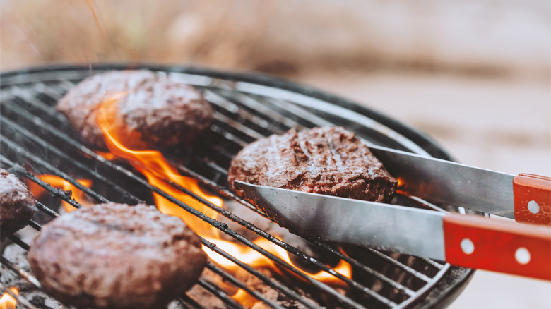 Close-up of tongs handling a plump burger on a hot grill in the foreground with two more burgers and flames behind.