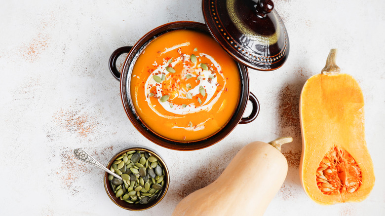 Pot of butternut squash soup next to cut squash and pepita topping