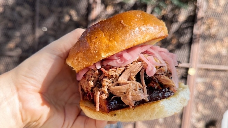 A hand holding a pulled pork sandwich with sausage and pickled onions from Lewis Barbecue