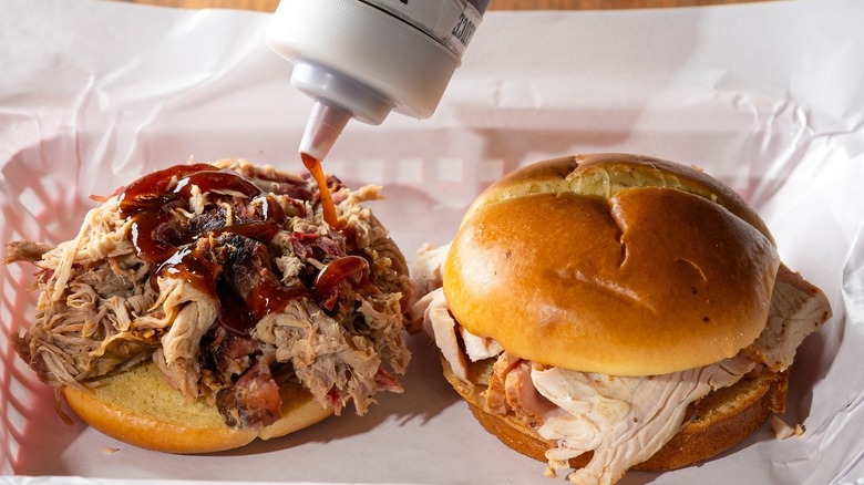 A person pouring sauce on a pulled pork sandwich next to a turkey sandwich from Pappy's Smokehouse