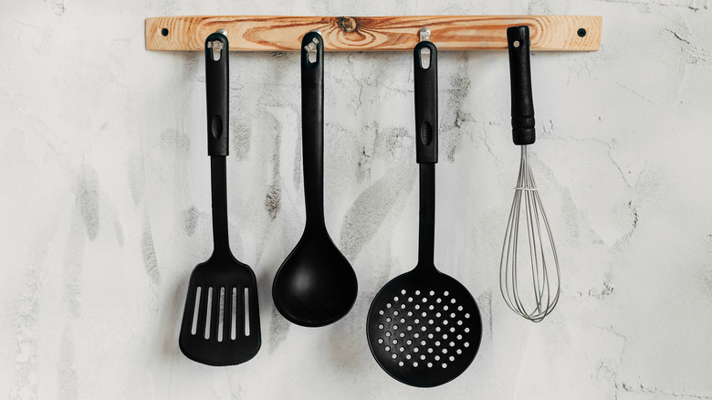 Black plastic and metal cooking tools hanging on a wooden rack.